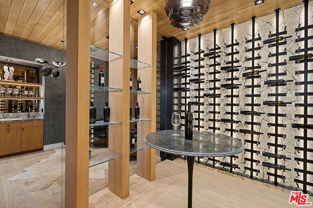 This sophisticated wine cellar features a dedicated wall-mounted metal racking system for wine storage and a central circular table for tasting. To the left, a glass-shelved display unit with light wood supports adds a modern touch, while a small wet bar area with cabinetry is visible in the background. The space is finished with light wood ceilings and flooring, creating a warm and refined atmosphere.
