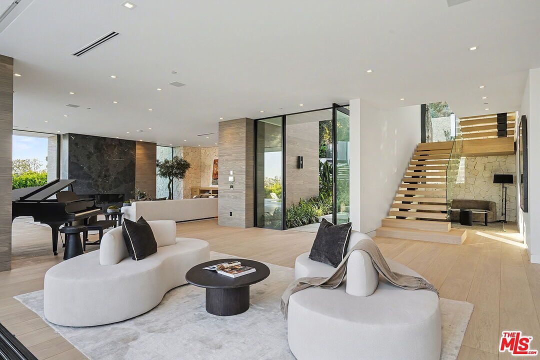 This expansive, open-concept living room features light wood flooring and a neutral color palette, creating a bright and airy atmosphere. The space is anchored by two curved, cream-colored sofas arranged around a dark circular coffee table, with a grand piano positioned near a dark stone-clad wall. A floating staircase with glass railings leads to the upper level, while large glass doors provide a seamless transition to the outdoor area.