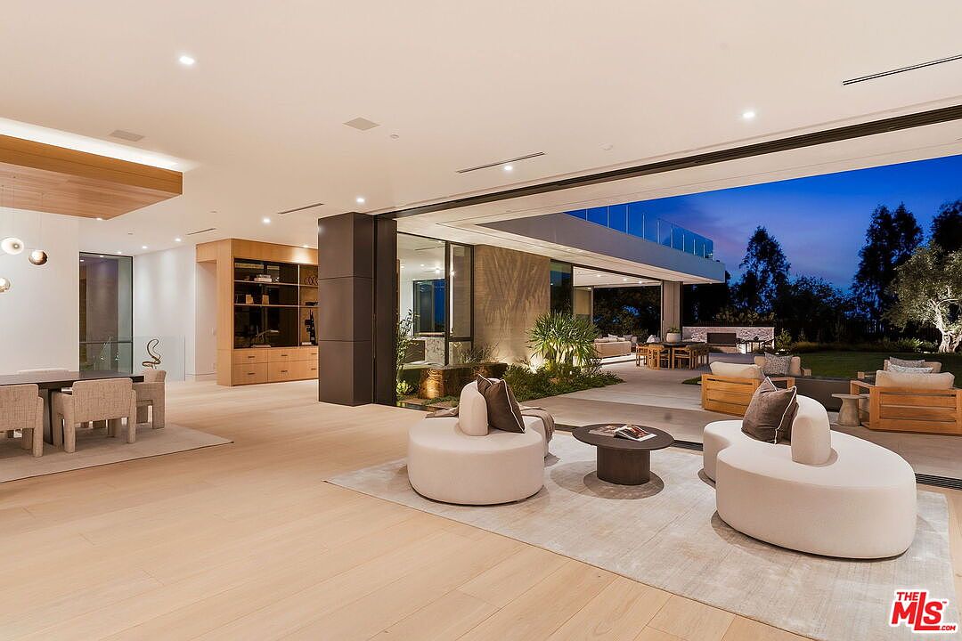 This expansive, open-concept living area features light wood flooring and a seamless transition to the outdoor patio through large, retractable glass walls. The space is furnished with modern, curved seating arrangements and a minimalist coffee table, creating a sophisticated and airy atmosphere. The interior flows effortlessly into a dining area on the left and a lush, illuminated outdoor garden space, emphasizing a luxurious indoor-outdoor lifestyle.