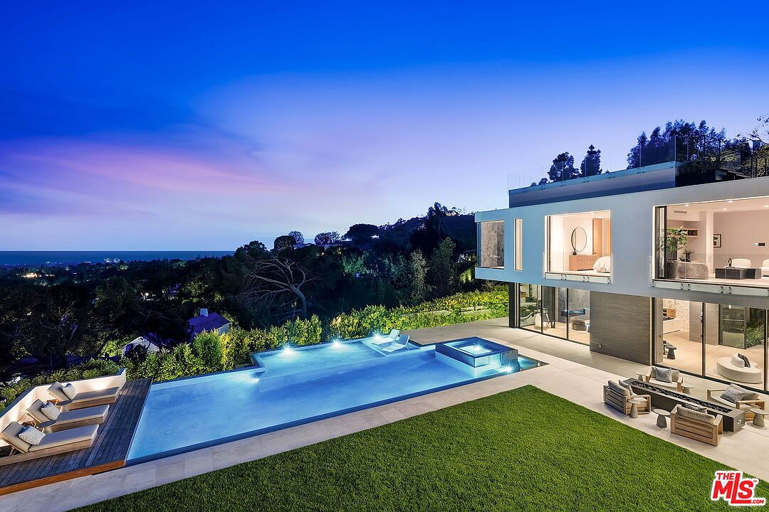 This modern luxury residence features a sleek, multi-level architectural design with expansive glass walls that seamlessly connect the indoor living spaces to the outdoors. The backyard is highlighted by a long, rectangular infinity-edge pool and integrated spa, set against a backdrop of lush hillside greenery and a distant ocean view. A manicured lawn and a wooden deck with lounge seating provide an inviting space for outdoor relaxation and entertaining at twilight.