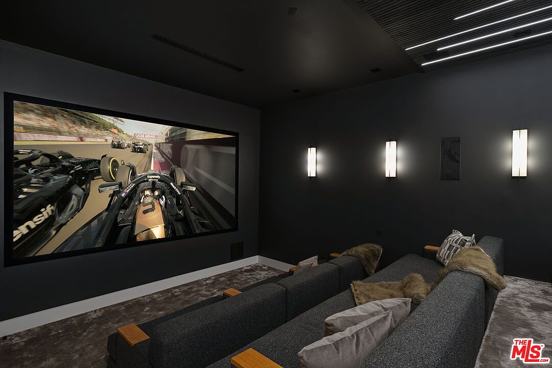 This dedicated home theater room features a large projection screen displaying a Formula 1 race, set against a dark, moody color palette. The space is furnished with tiered rows of plush, dark gray seating accented with textured throw pillows and faux-fur blankets. Wall-mounted sconces provide soft, ambient lighting, while the ceiling incorporates linear lighting elements to enhance the cinematic atmosphere.