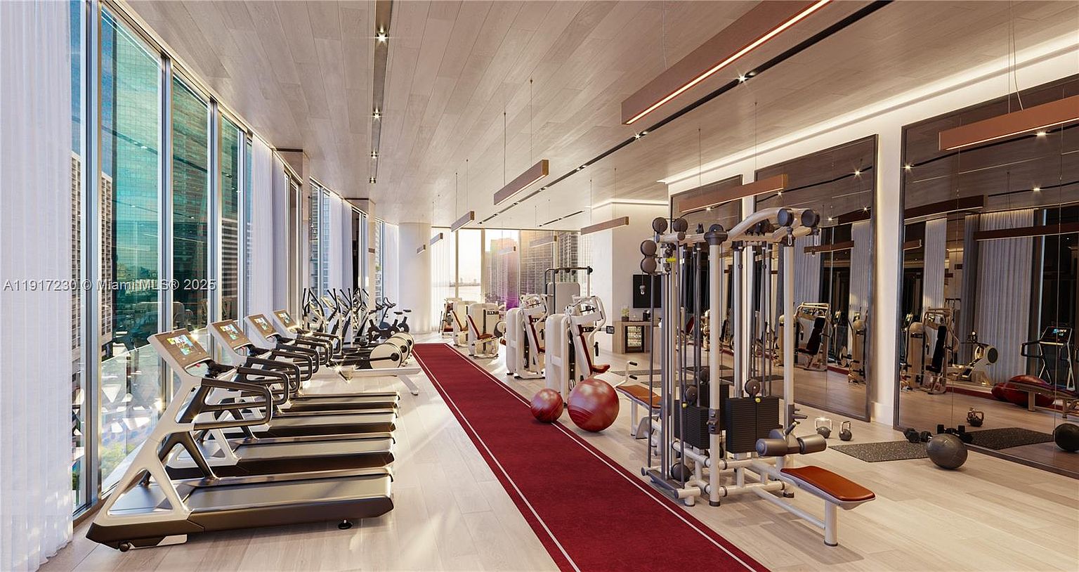 This modern, high-end fitness center features a long row of treadmills positioned alongside expansive floor-to-ceiling windows that offer an abundance of natural light. The space is equipped with professional-grade strength training machines, exercise balls, and a central red runner rug that adds a vibrant pop of color to the neutral wood-toned interior. The room is designed with a sleek, minimalist aesthetic, utilizing mirrored walls to enhance the sense of depth and openness.