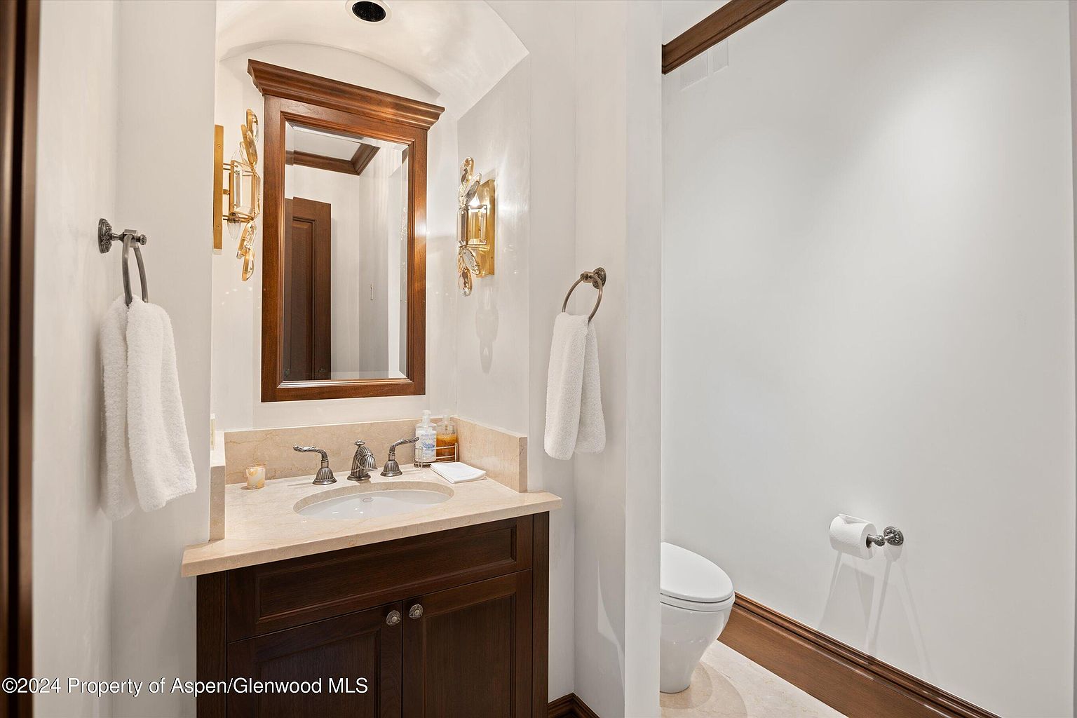 This is a well-appointed guest bathroom featuring a dark wood vanity with a light-colored countertop and an oval sink. A framed mirror hangs above the vanity, flanked by decorative sconces. A toilet is visible to the right, and the walls are painted in a neutral tone, creating a clean and inviting space.