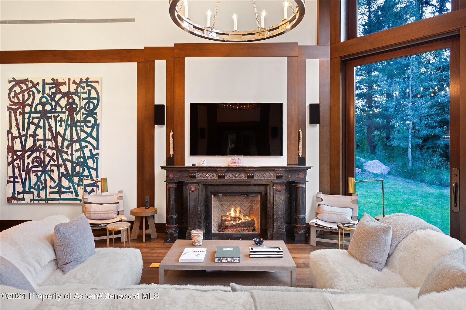 This is an interior shot of a luxurious living room featuring a grand fireplace with a mounted television above it. The room is decorated with modern art, comfortable seating, and wooden architectural details, creating a warm and inviting atmosphere. A large window offers a view of the outdoor landscape, enhancing the room's connection to nature.