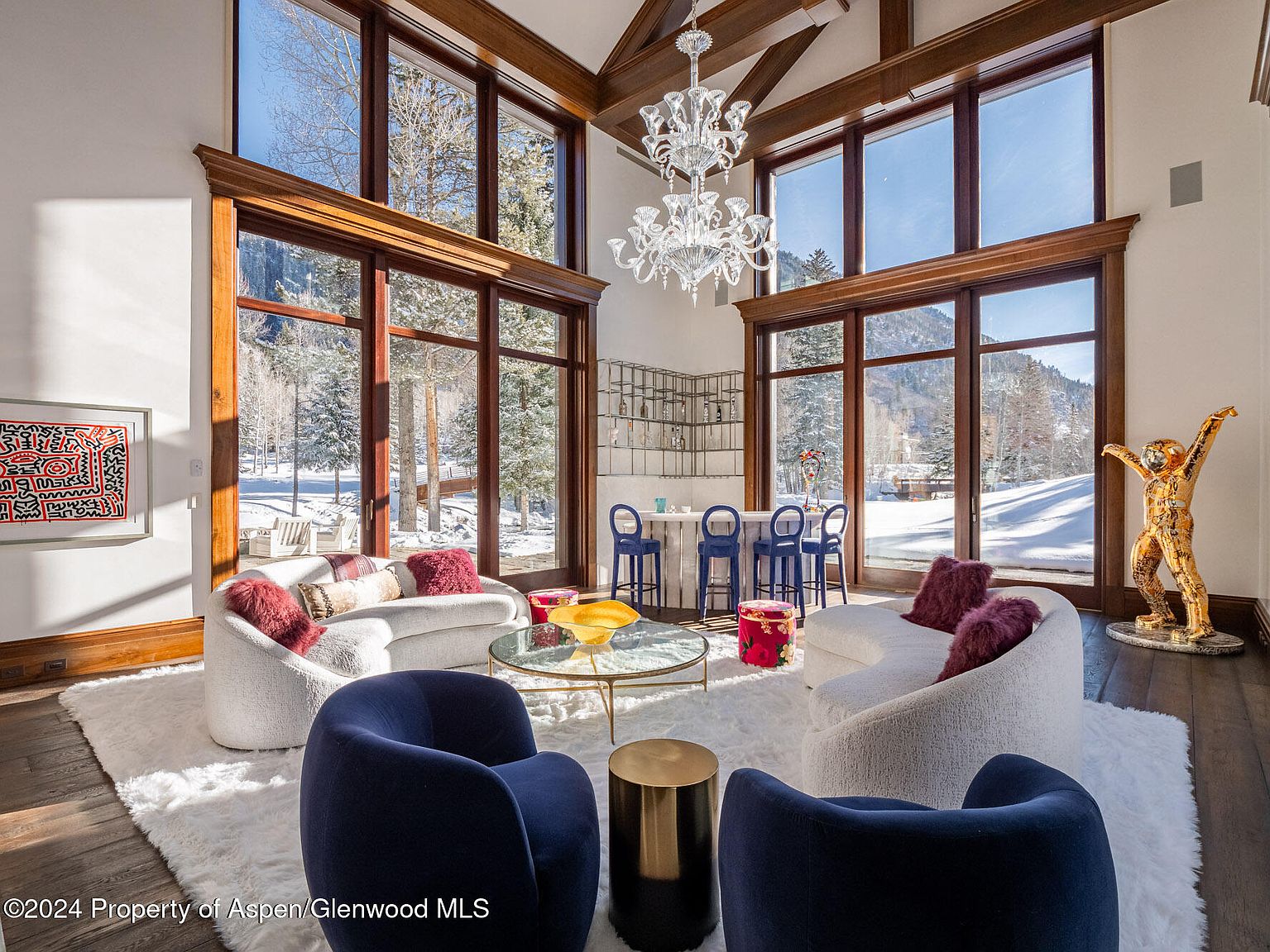 This is a luxurious living room featuring floor-to-ceiling windows that offer a stunning view of a snow-covered landscape. The room is furnished with curved sofas, plush armchairs, and a unique chandelier, creating an elegant and inviting atmosphere. The hardwood floors and area rug add warmth and texture to the space.