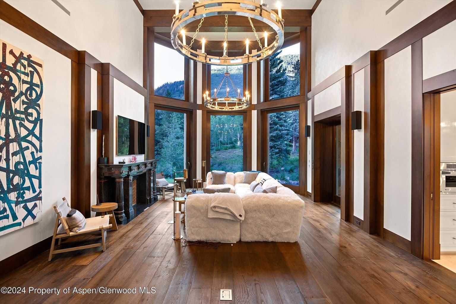 This is a spacious living room featuring hardwood floors, a large fur-like sectional sofa, and a striking chandelier. The room is framed by large windows offering a view of the outdoors, and the walls are accented with dark wood trim. A fireplace and artwork add to the room's luxurious and inviting atmosphere.