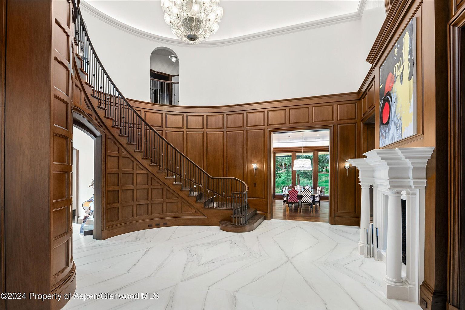This grand foyer showcases a curved staircase with dark wood and wrought iron railings, complemented by rich wood paneling and a striking chandelier. The white marble flooring with geometric patterns adds a touch of elegance, while an adjacent dining room is visible through an open doorway. The overall impression is one of luxury and sophisticated design.