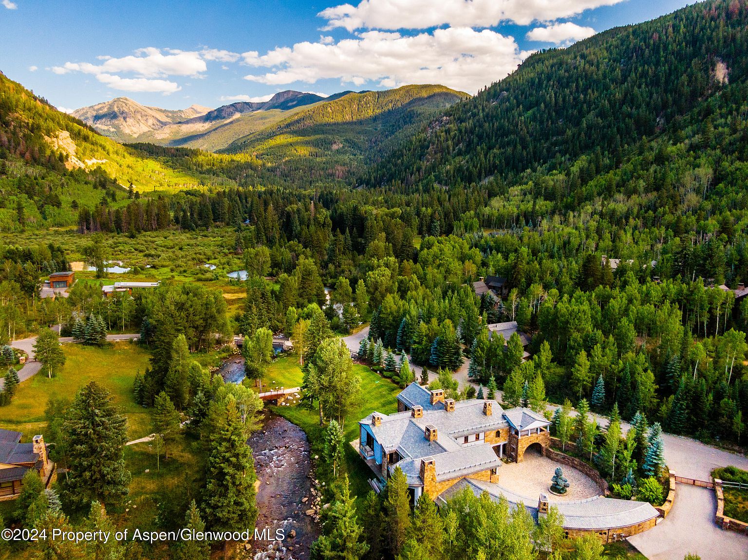 This aerial view showcases a luxurious estate nestled in a lush, mountainous landscape. A winding river flows alongside the property, enhancing its natural beauty. The home features a stone exterior, a complex roofline, and a grand entrance, exuding elegance and privacy.