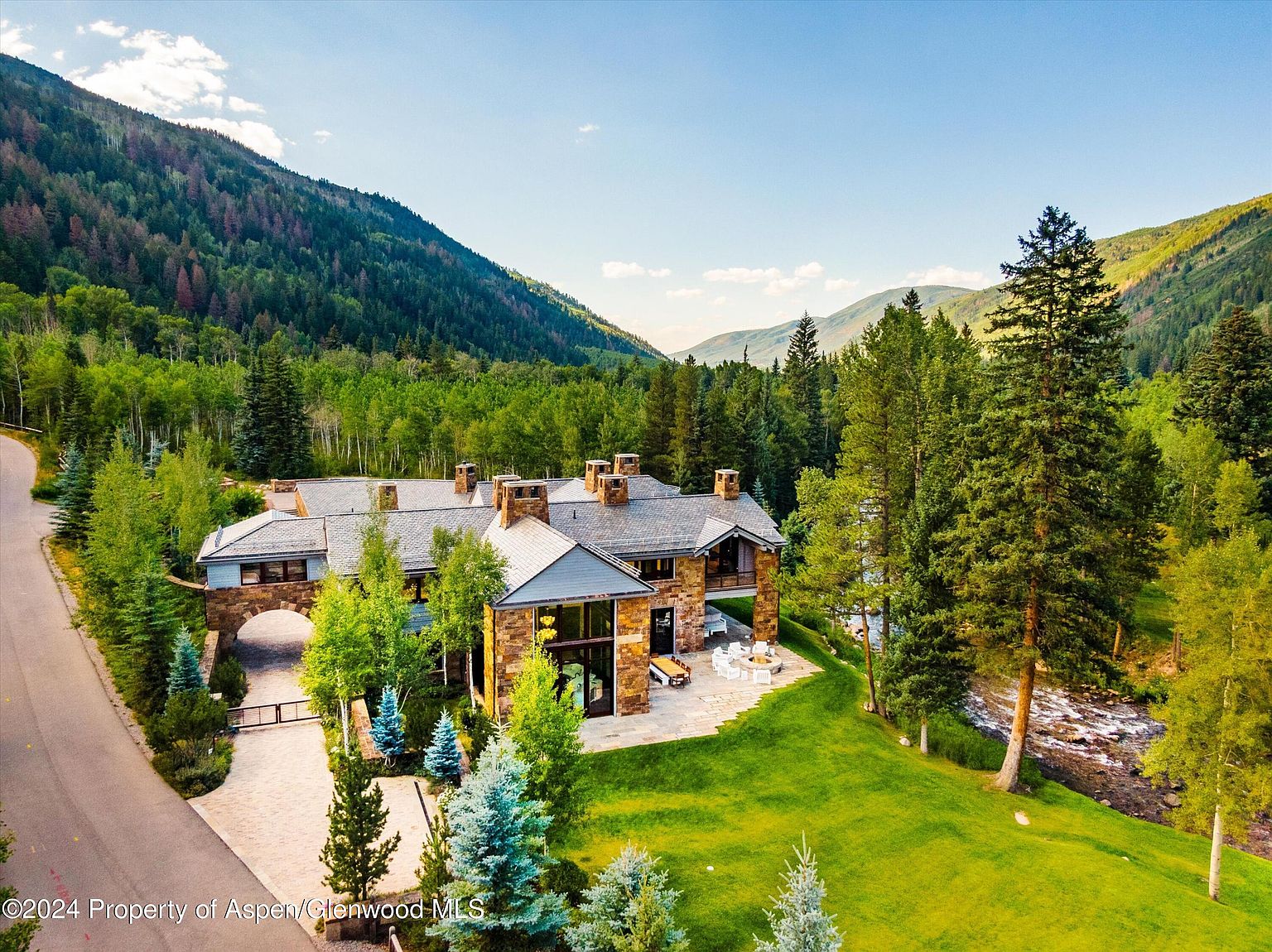 This aerial view showcases a luxurious mountain estate nestled amidst lush greenery and towering trees. The stone and timber home features a grand entrance archway, multiple chimneys, and a sprawling lawn that extends to a gently flowing river. The overall impression is one of secluded elegance and natural beauty, perfect for a discerning buyer seeking a private retreat.