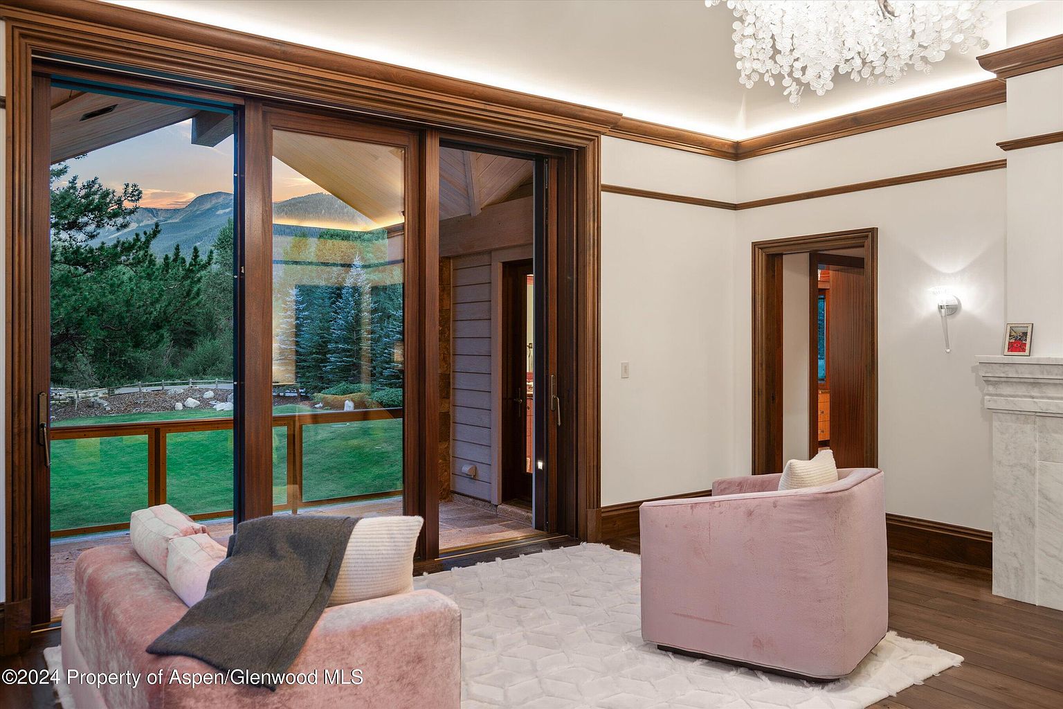 This interior shot showcases a luxurious living room with a blend of modern and classic design elements. The room features a large sliding glass door offering a view of the outdoors, complemented by rich wood trim and a decorative chandelier. Plush pink seating and a light-colored rug add a touch of elegance and comfort to the space.