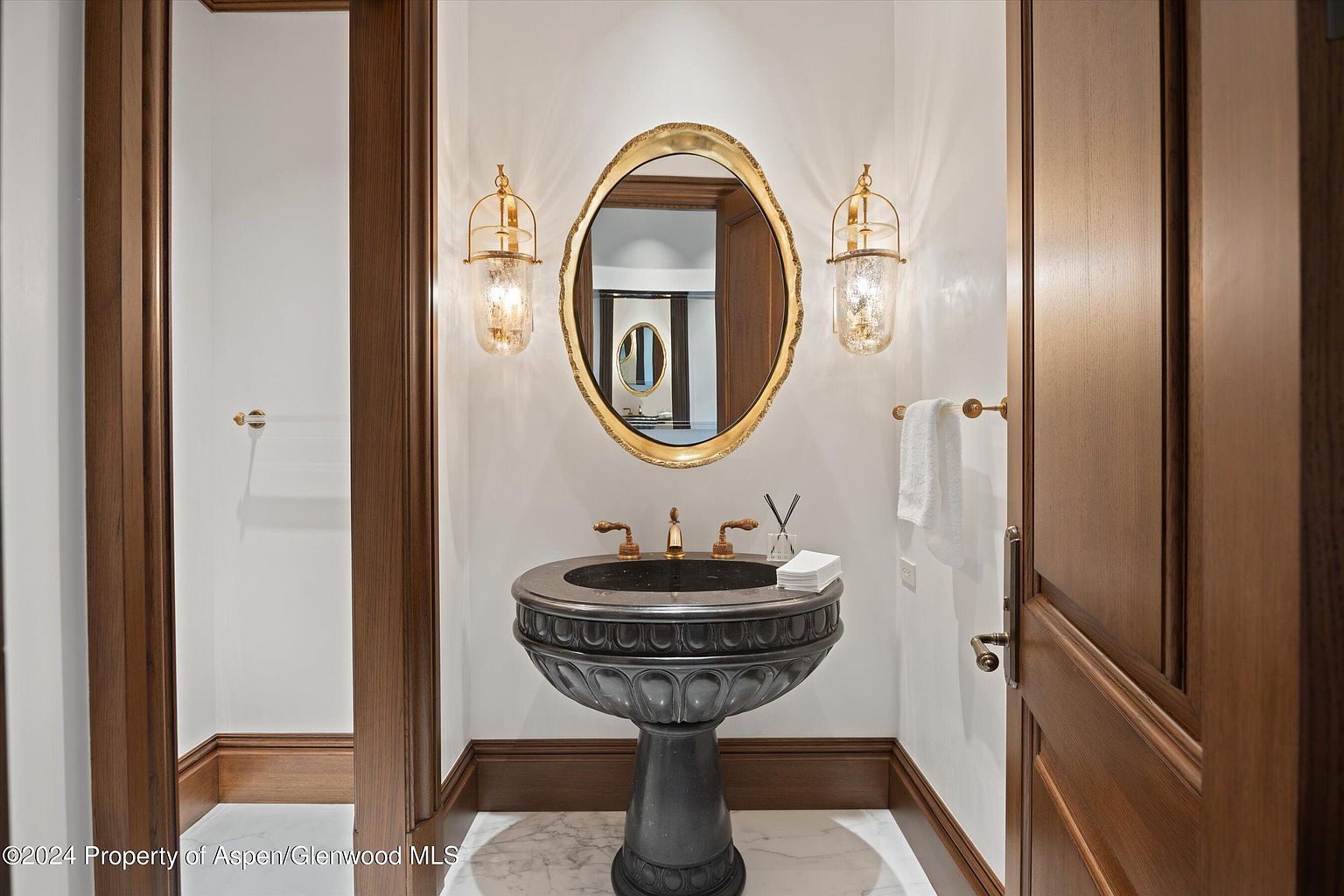 This is a well-appointed guest bathroom featuring a pedestal sink with a dark stone basin and gold fixtures. An ornate gold-framed mirror hangs above the sink, flanked by sconces that provide warm lighting. The room is finished with wood trim and a marble floor, creating a luxurious and elegant space.