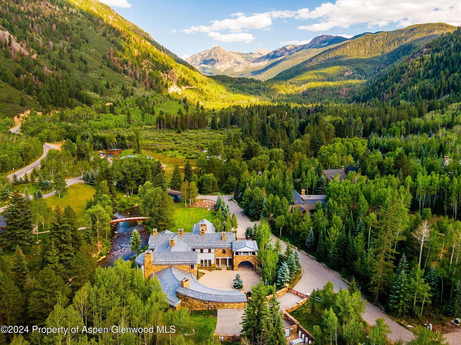 This aerial view showcases a luxurious estate nestled in a lush, mountainous landscape. The property features a grand stone mansion with a circular driveway, multiple chimneys, and a meticulously landscaped yard. A river flows nearby, crossed by a charming bridge, adding to the serene and private setting.