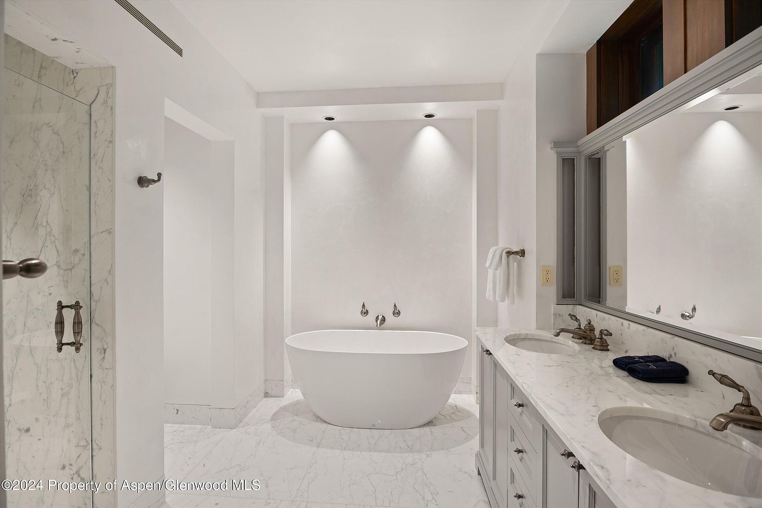 This is a bright and luxurious primary bathroom featuring a freestanding white bathtub as a focal point. The room is decorated in a clean, modern style with white walls and marble flooring. A double vanity with marble countertops and bronze fixtures adds elegance, while a glass-enclosed shower is visible to the left.