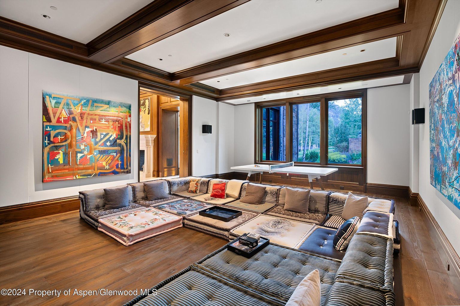 This interior shot showcases a unique living room featuring a modular floor-level seating arrangement with various cushions and patterns. The room is accented by dark wood trim, hardwood floors, and large abstract art pieces on the walls. A ping pong table sits near the windows, which offer a view of the outdoors.