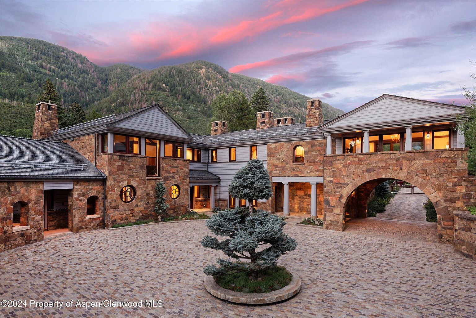 This is a stunning front view of a luxurious stone house with a grand driveway. The architecture features a blend of stone and siding, with arched entryways and well-lit windows creating a warm and inviting atmosphere. The property is nestled against a backdrop of lush, green mountains under a vibrant sunset sky, enhancing its appeal and sense of exclusivity.