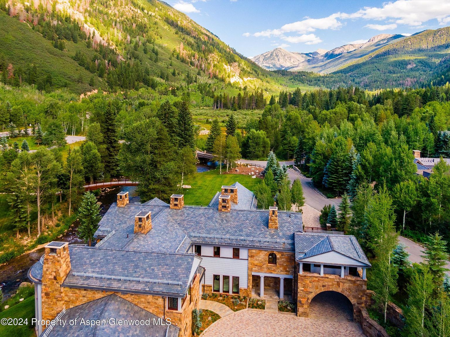 This aerial view showcases a luxurious estate nestled in a lush, mountainous landscape. The home features a stone exterior, a slate roof with multiple chimneys, and a grand arched entryway. The property is surrounded by mature trees, a winding river with bridges, and manicured lawns, creating a serene and private setting.