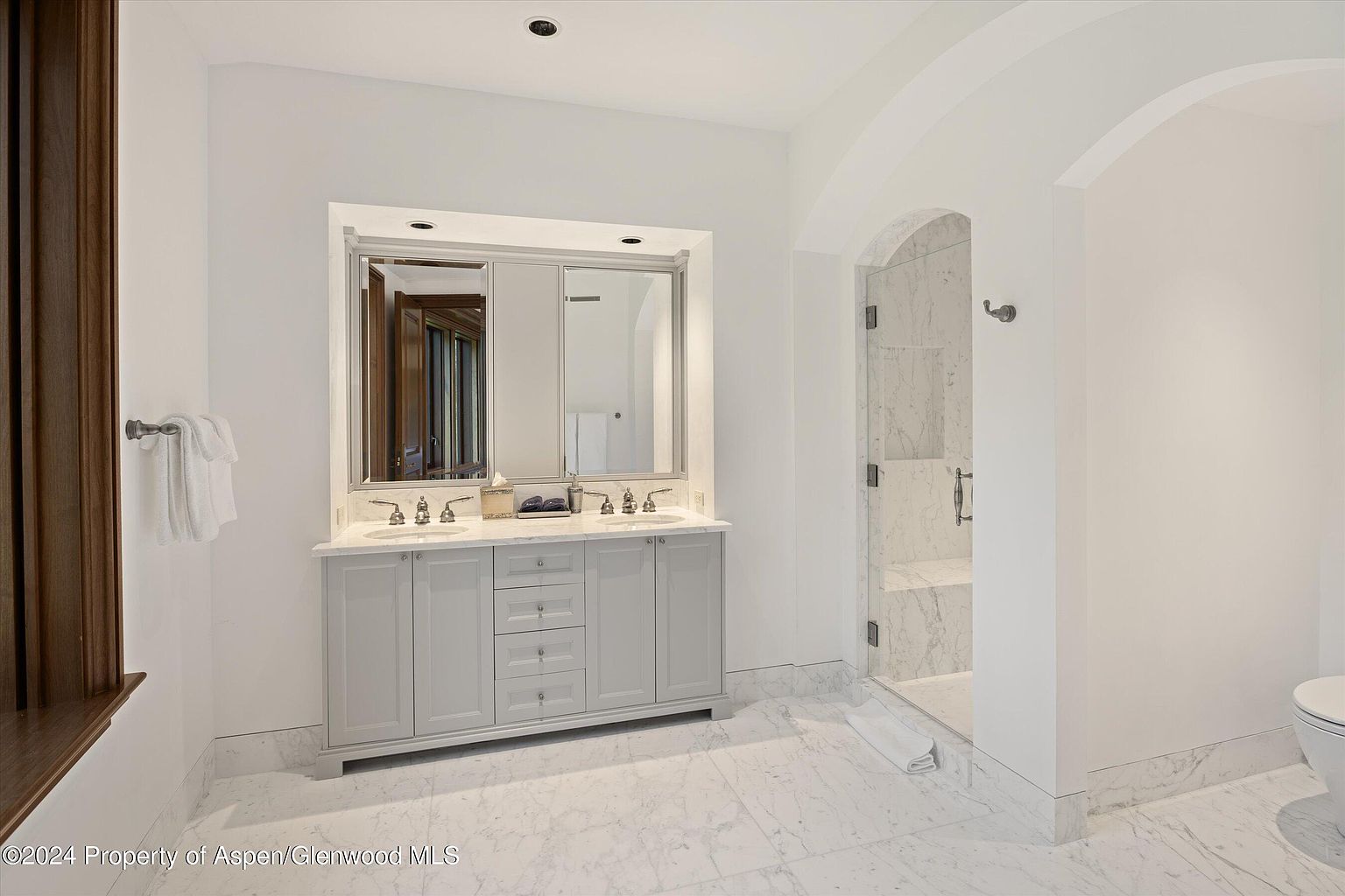 This is a bright and elegant primary bathroom featuring a double vanity with a light gray cabinet, marble countertops, and large mirrors. The bathroom has marble flooring and an arched doorway leading to a glass-enclosed shower. The overall impression is clean, luxurious, and well-maintained.