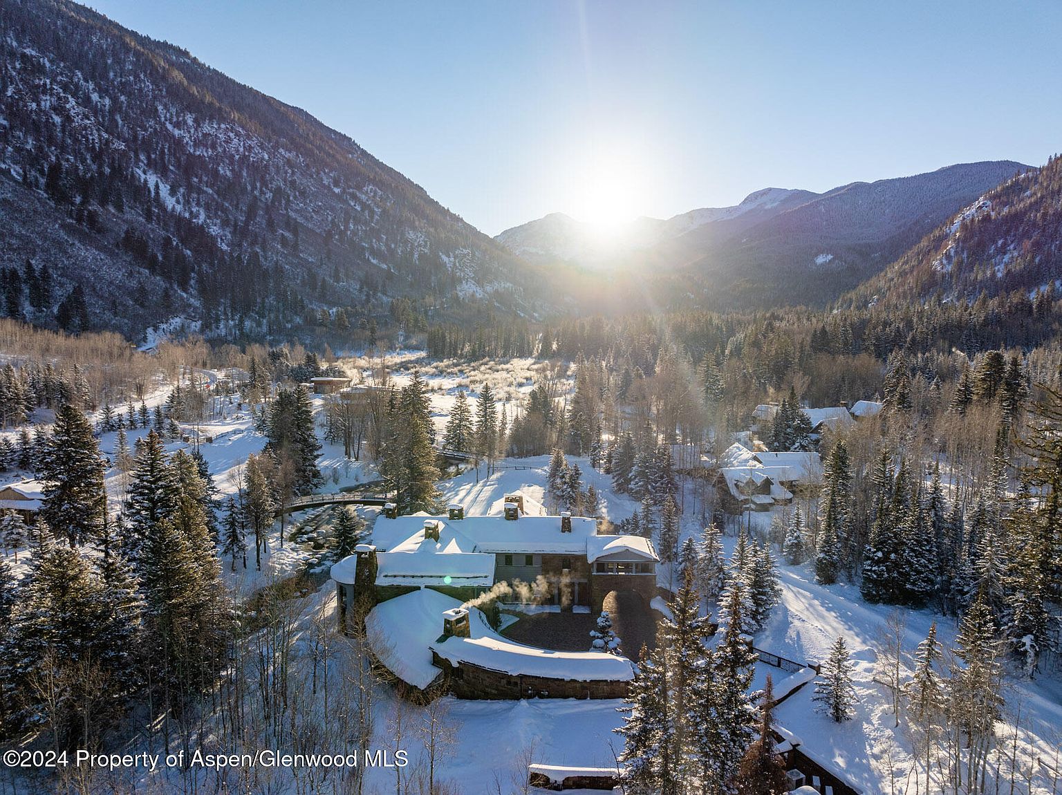 This aerial view showcases a luxurious mountain home nestled in a snow-covered valley. The property features a stone exterior, multiple chimneys, and a curved stone wall, blending seamlessly with the natural landscape. Towering pine trees and snow-capped mountains create a serene and picturesque setting, emphasizing the property's privacy and exclusivity.