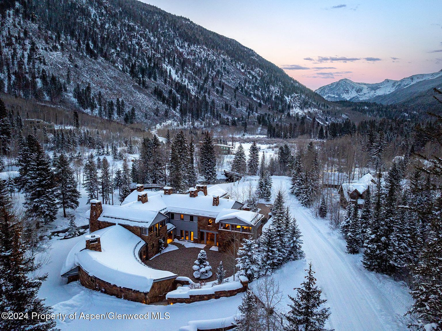 This aerial view showcases a luxurious mountain home nestled in a snowy landscape. The property features a stone and gray exterior, multiple chimneys, and a circular driveway. Towering snow-covered trees and a majestic mountain backdrop enhance the sense of privacy and natural beauty, making it an ideal retreat.