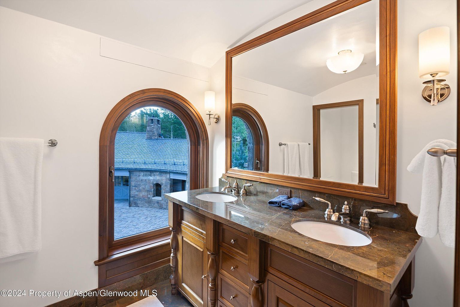 This is a primary bathroom featuring a double vanity with a dark marble countertop and wooden cabinetry. An arched window provides natural light and a view of the exterior, while a large framed mirror reflects the interior space, including a glimpse of another room. The overall impression is luxurious and well-appointed.