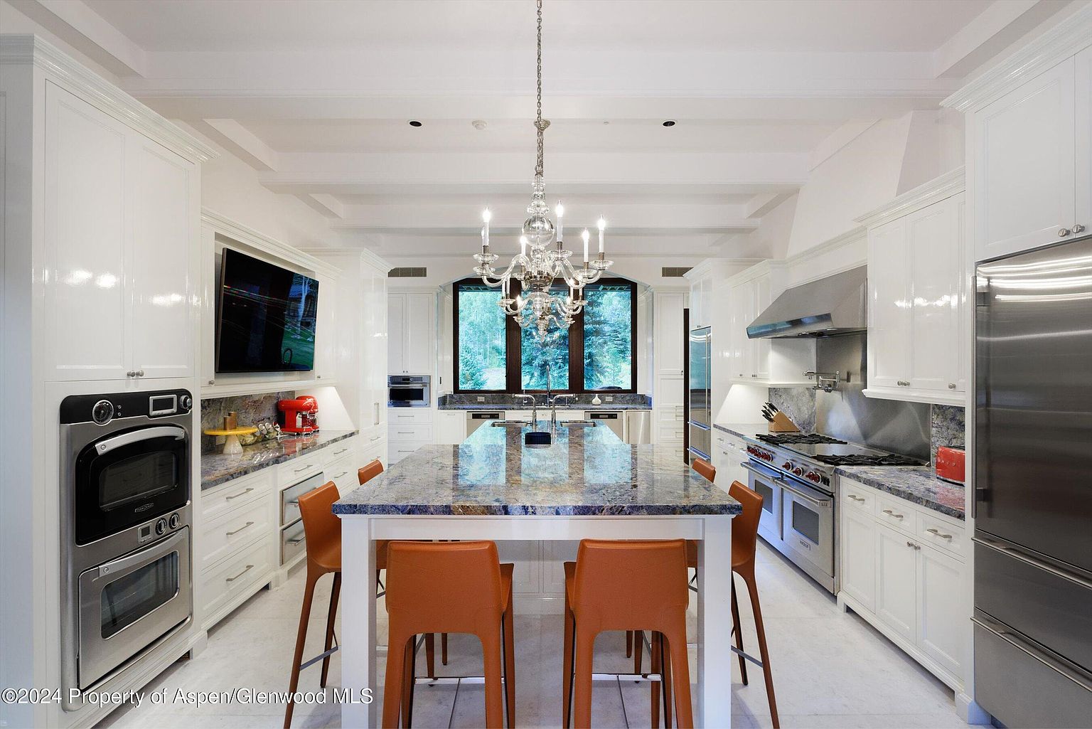 This is a bright and spacious kitchen featuring white cabinetry, stainless steel appliances, and a large island with a unique granite countertop. A chandelier hangs above the island, and a large window provides natural light and a view of the outdoors. The perspective is a wide shot, emphasizing the kitchen's length and the symmetry of its design.