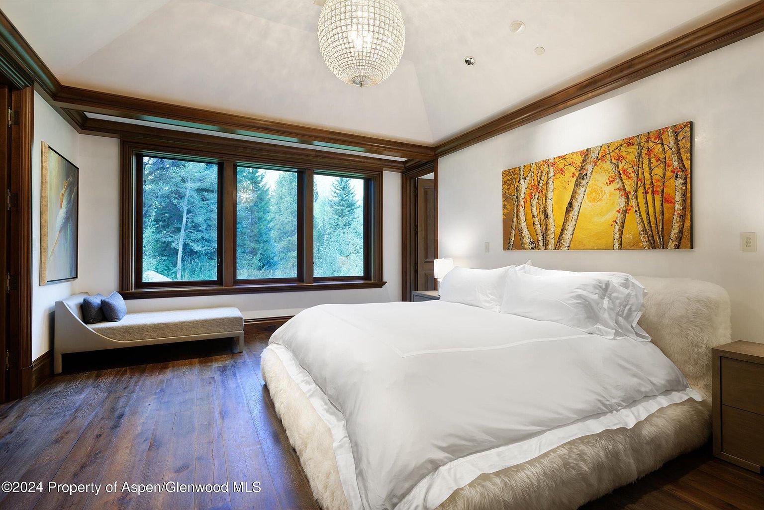 This is a primary bedroom featuring a large bed with white linens and a fur-like base, a window with a view of trees, and an abstract painting above the bed. The room has hardwood floors and wood trim, with a chandelier hanging from the ceiling. The overall impression is luxurious and serene.
