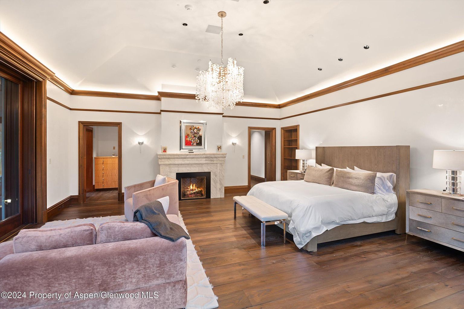 This is a spacious primary bedroom featuring hardwood floors, a fireplace, and a large bed with a neutral-toned headboard. The room is well-lit with a chandelier and recessed lighting, complemented by natural light from a window. A seating area with a plush sofa adds to the luxurious feel of the space.