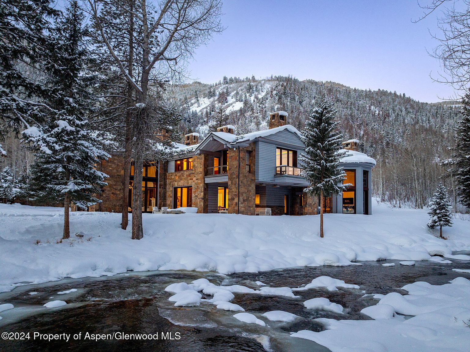 This is a stunning front view of a luxurious mountain home in winter. The house features a combination of stone and siding, with large windows offering views of the interior lighting. A partially frozen river flows in the foreground, adding to the serene and picturesque setting, while snow-covered mountains rise in the background.