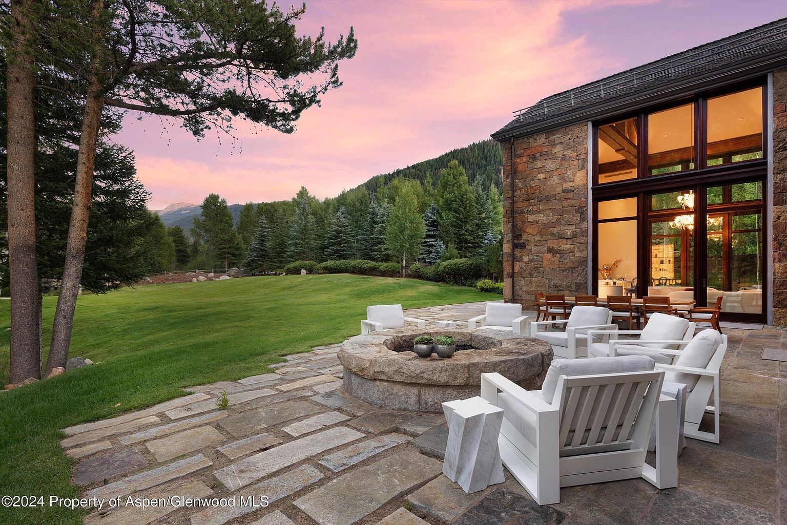 This image showcases a luxurious outdoor patio area featuring a stone fire pit surrounded by modern white lounge chairs. The patio is paved with large stone tiles, and a well-manicured lawn extends towards a backdrop of lush green trees and a mountain range under a pink-tinged sky. The stone exterior of the adjacent building adds a touch of rustic elegance to the scene.