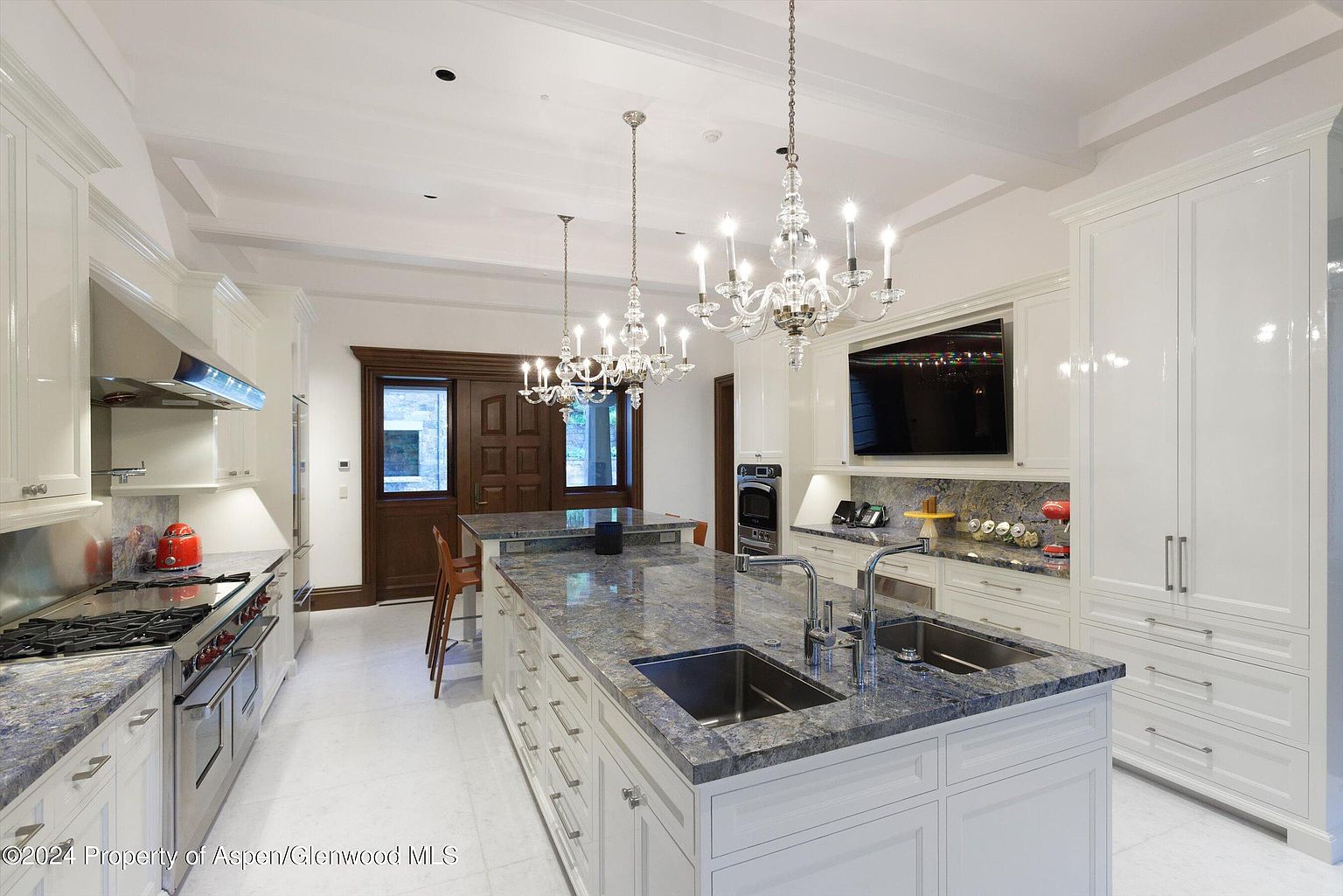 This is a bright and luxurious kitchen featuring white cabinetry, granite countertops, and stainless steel appliances. The kitchen includes a large island with a sink and ample storage, illuminated by elegant chandeliers. A television is mounted above the countertop, and a doorway is visible in the background, offering a glimpse into another room.