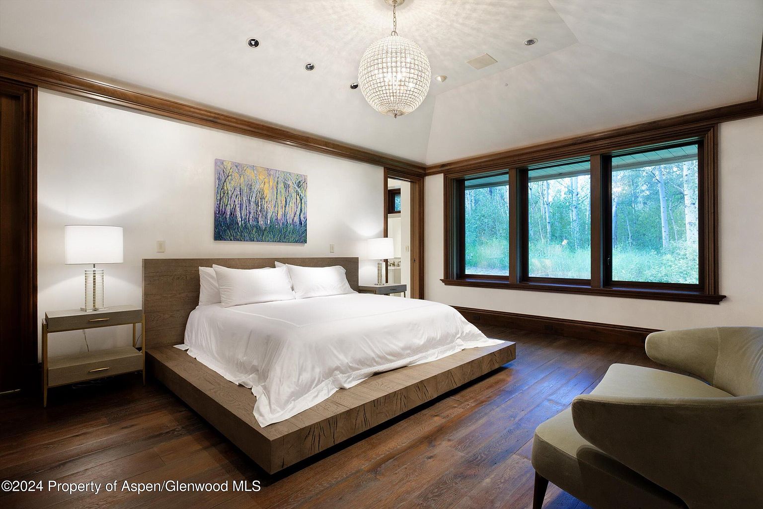 This is a primary bedroom featuring a large platform bed with a white duvet and pillows, flanked by nightstands with lamps. The room has dark wood floors and trim, with a large window offering a view of the trees outside. A modern chandelier hangs from the ceiling, and a piece of artwork is displayed above the bed, creating a luxurious and serene atmosphere.
