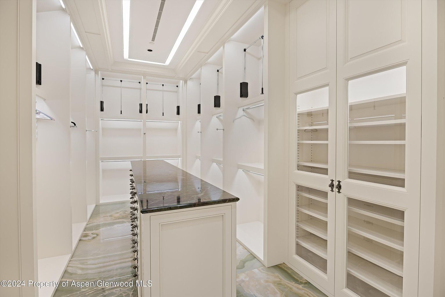 This is a well-organized walk-in closet featuring custom white cabinetry, shelving, and hanging rods. A central island with a dark countertop provides additional storage and workspace. The closet is illuminated by recessed lighting and has a luxurious feel, suggesting ample space for clothing and accessories.