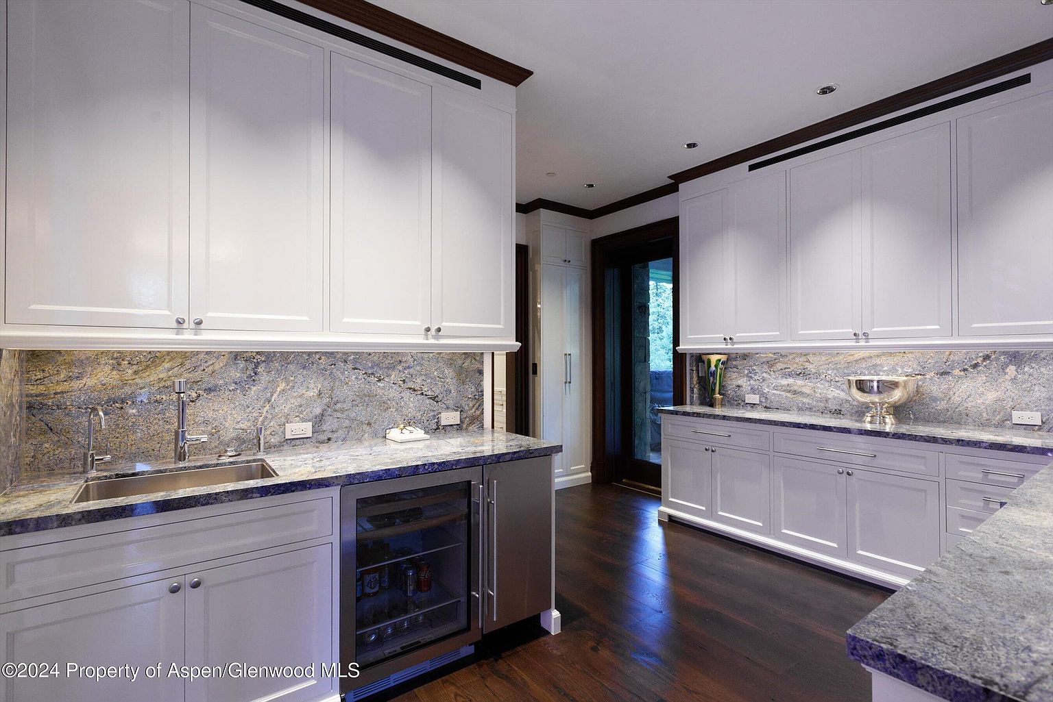This interior shot showcases a well-appointed kitchen with white cabinetry and a striking granite backsplash and countertops. A stainless steel sink and beverage cooler are visible, adding to the kitchen's functionality. The dark wood flooring provides a rich contrast to the light cabinetry, enhancing the room's elegance.
