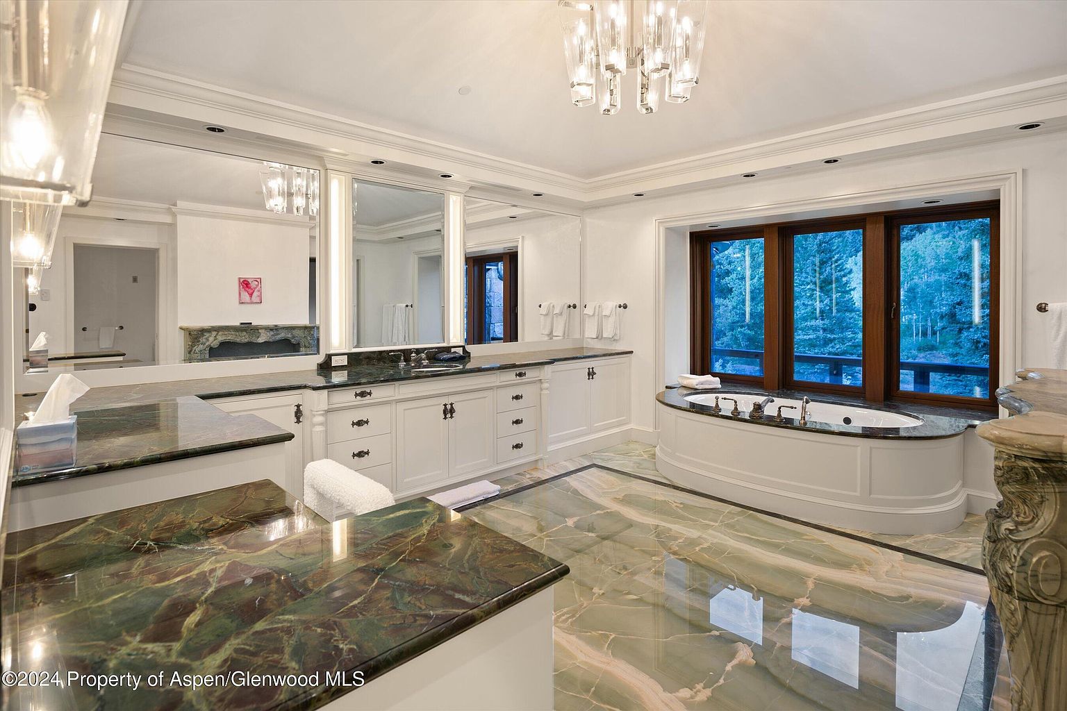 This is a luxurious primary bathroom featuring a large vanity with a green marble countertop, white cabinetry, and a large mirror. A soaking tub is positioned in front of a large window offering a view of the outdoors. The floor is also covered in green marble, adding to the opulent feel of the space.