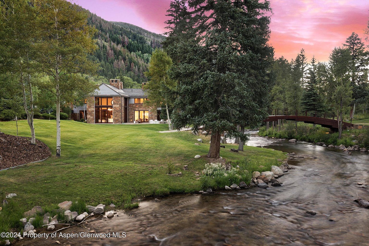 This exterior shot showcases the rear of a luxurious stone house with large windows overlooking a well-manicured lawn and a flowing river. A charming wooden bridge spans the river, adding to the property's appeal. The scene is bathed in the warm hues of a sunset, creating a serene and inviting atmosphere.
