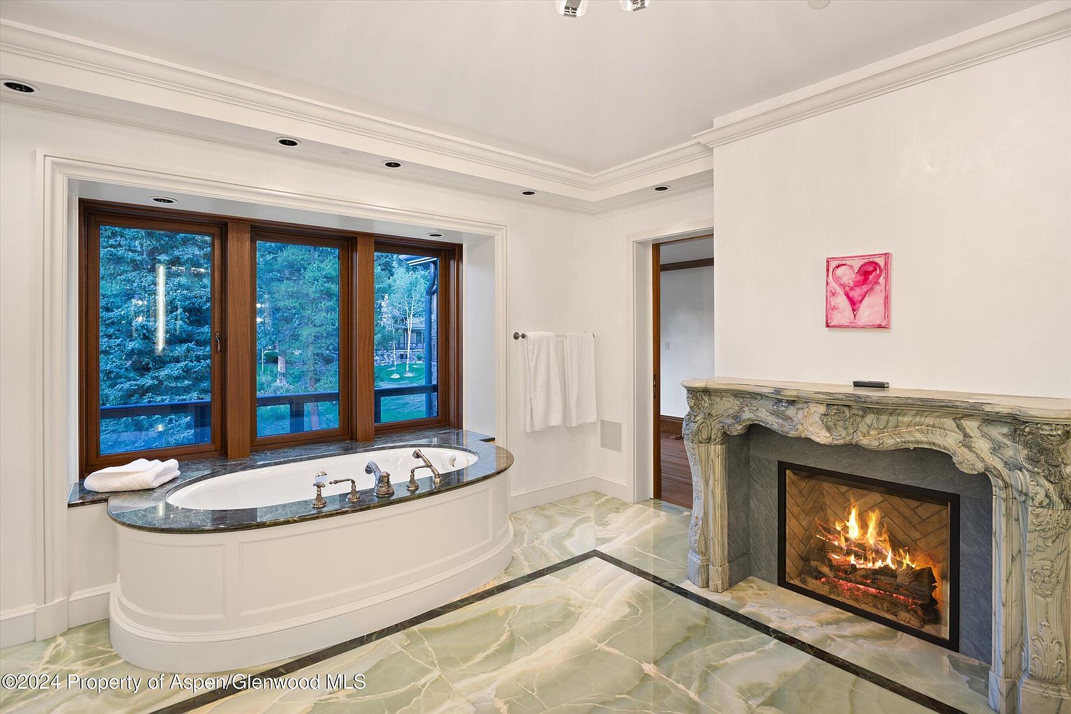This is a luxurious primary bathroom featuring a large oval soaking tub positioned beneath a window offering a view of the outdoors. The bathroom boasts a marble fireplace, adding a touch of elegance and warmth. The floor is covered in green marble with black accents, complementing the overall sophisticated design.