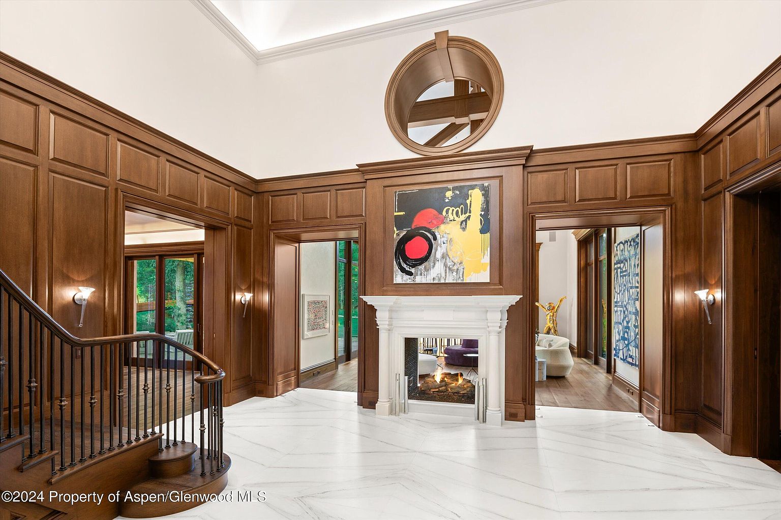 This grand foyer features rich wood paneling, a striking marble floor with a geometric pattern, and a staircase with wrought iron railings. A large fireplace with a white mantel is centered on the wall, topped with an abstract painting and a circular architectural detail above. The space is well-lit and exudes a sense of luxury and sophistication.