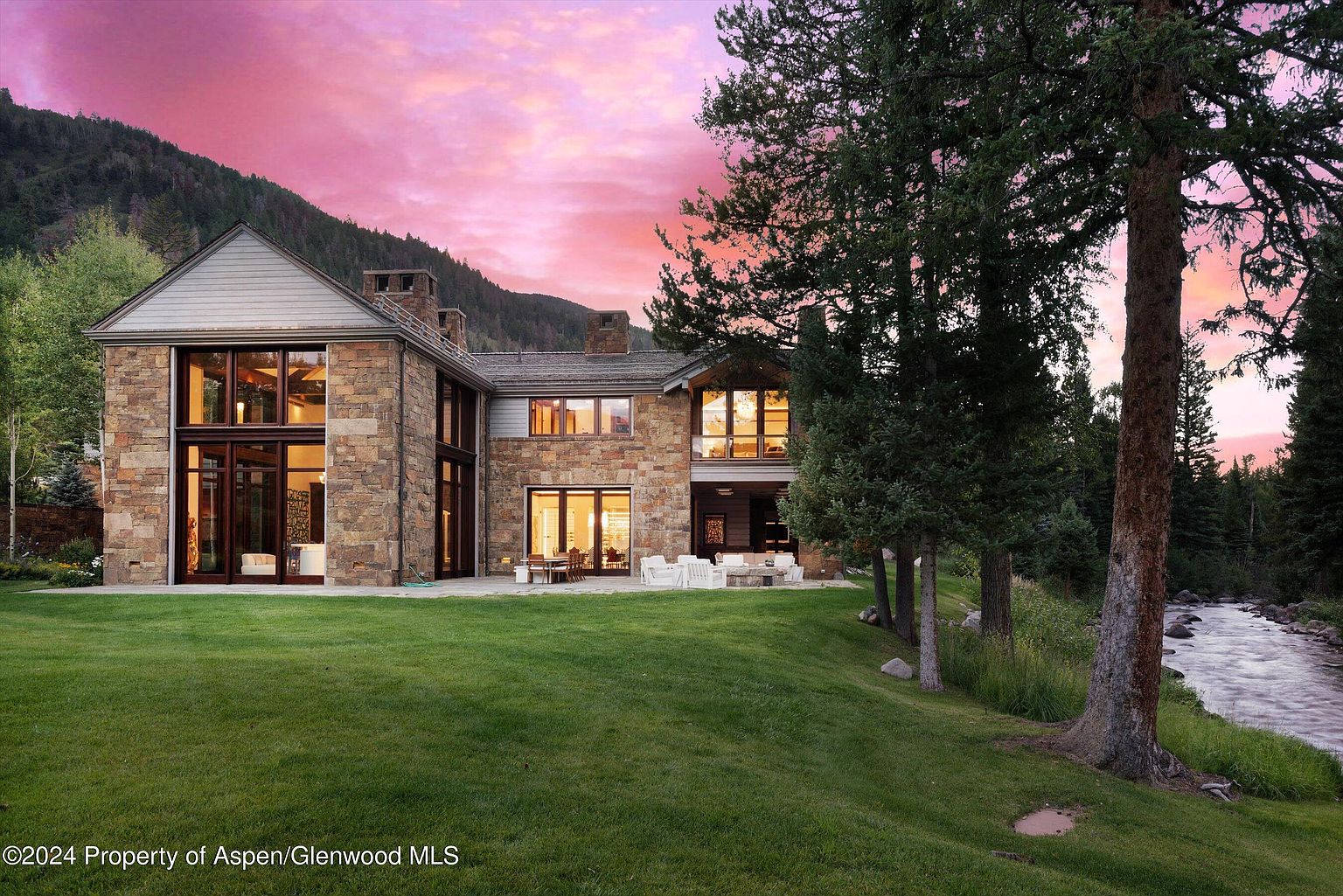 This image showcases the rear exterior of a luxurious stone house with large windows offering views of the interior. A well-manicured lawn extends from the house to a riverbank, with mature trees framing the scene. The sky is painted with vibrant pink hues, creating a serene and upscale ambiance.
