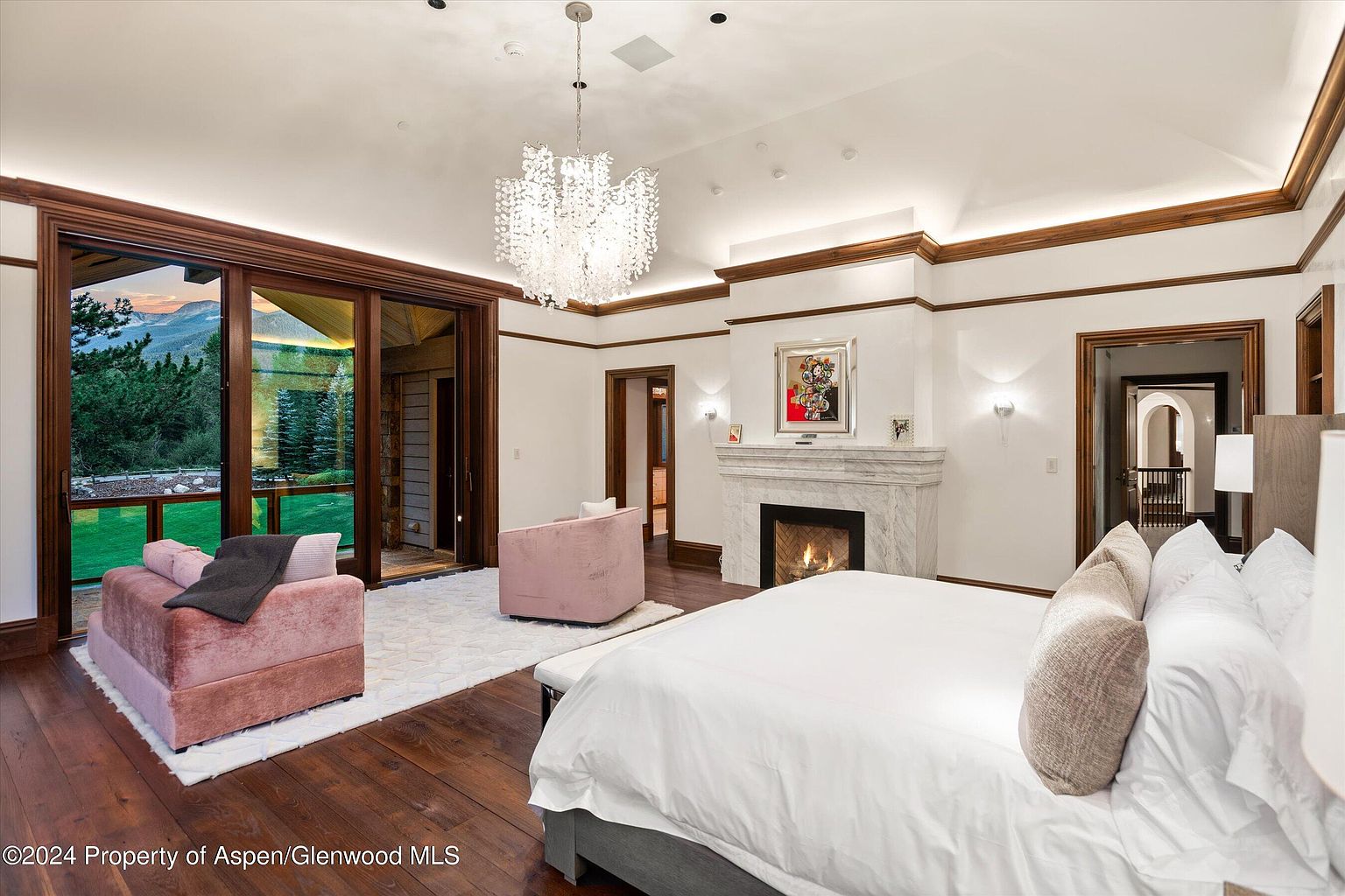 This is a luxurious primary bedroom featuring a king-size bed with white linens and neutral-toned pillows. The room boasts a marble fireplace, a modern chandelier, and large sliding glass doors that lead to an outdoor area with lush greenery. The dark hardwood floors and elegant trim add to the room's sophisticated ambiance.