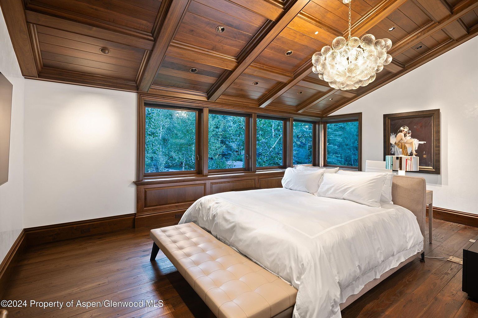 This is a primary bedroom featuring a large bed with white linens, a tufted bench at the foot of the bed, and a wall of windows overlooking a lush green landscape. The room has a warm and inviting feel, with rich wood flooring and a coffered wood ceiling adding architectural interest. A modern chandelier hangs above the bed, and artwork is displayed on the wall.