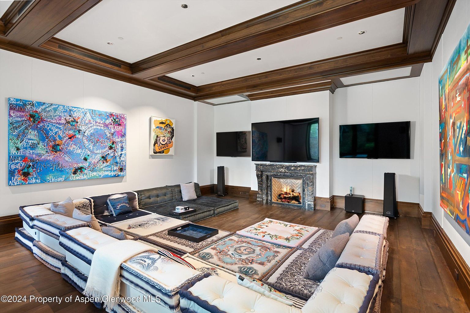 This interior shot showcases a luxurious living room with a unique, modular sofa arrangement, hardwood floors, and a coffered ceiling with wooden beams. The room features multiple large-screen televisions and a fireplace with a marble surround, complemented by colorful abstract art pieces on the walls, creating a modern and artistic ambiance.