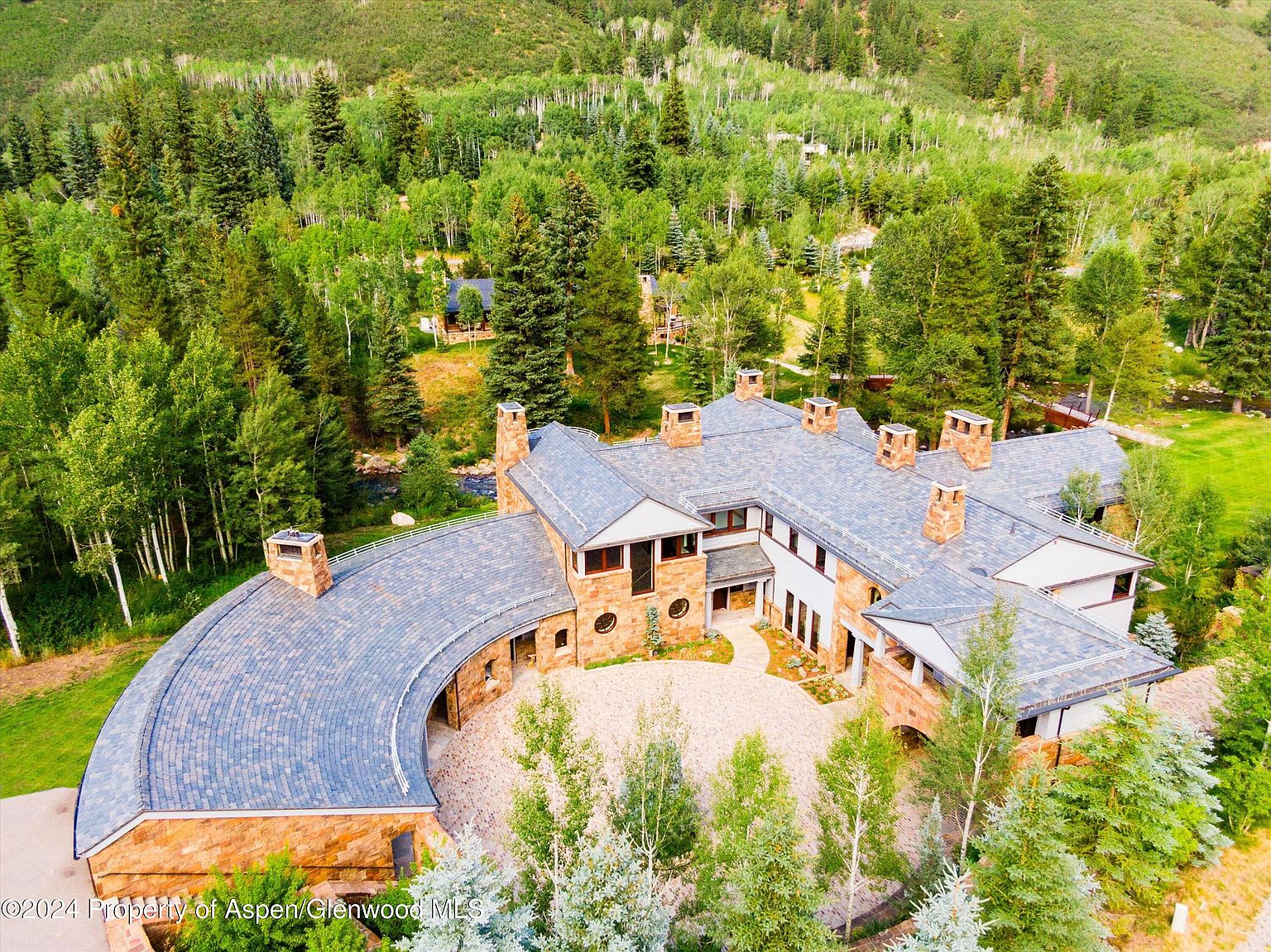 This aerial view showcases a luxurious estate nestled amidst lush greenery. The property features a unique, curved architectural design with stone and slate roofing, multiple chimneys, and a spacious courtyard. The surrounding landscape includes mature trees and manicured lawns, creating a serene and private setting.