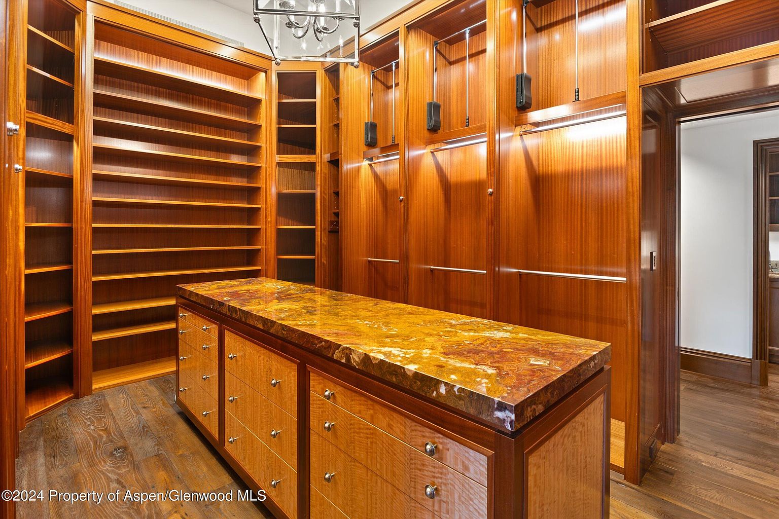 This is a well-organized walk-in closet featuring custom wood cabinetry and shelving. A central island with a marble countertop provides additional storage and a surface for organizing clothes. The closet is spacious and well-lit, offering a luxurious and functional space for clothing and accessories.
