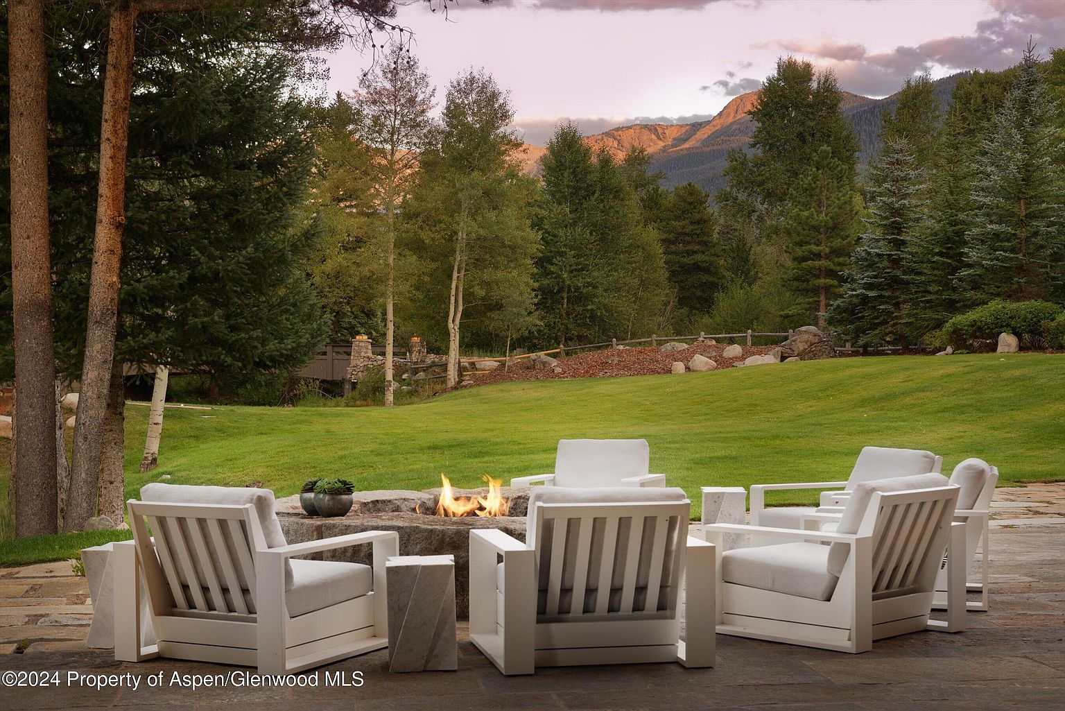 This image showcases a beautifully landscaped backyard featuring a stone patio with modern white outdoor furniture arranged around a fire pit. The lush green lawn extends towards a backdrop of mature trees and mountains, creating a serene and inviting outdoor living space. The scene is captured at dusk, with a soft, warm light enhancing the natural beauty of the setting.