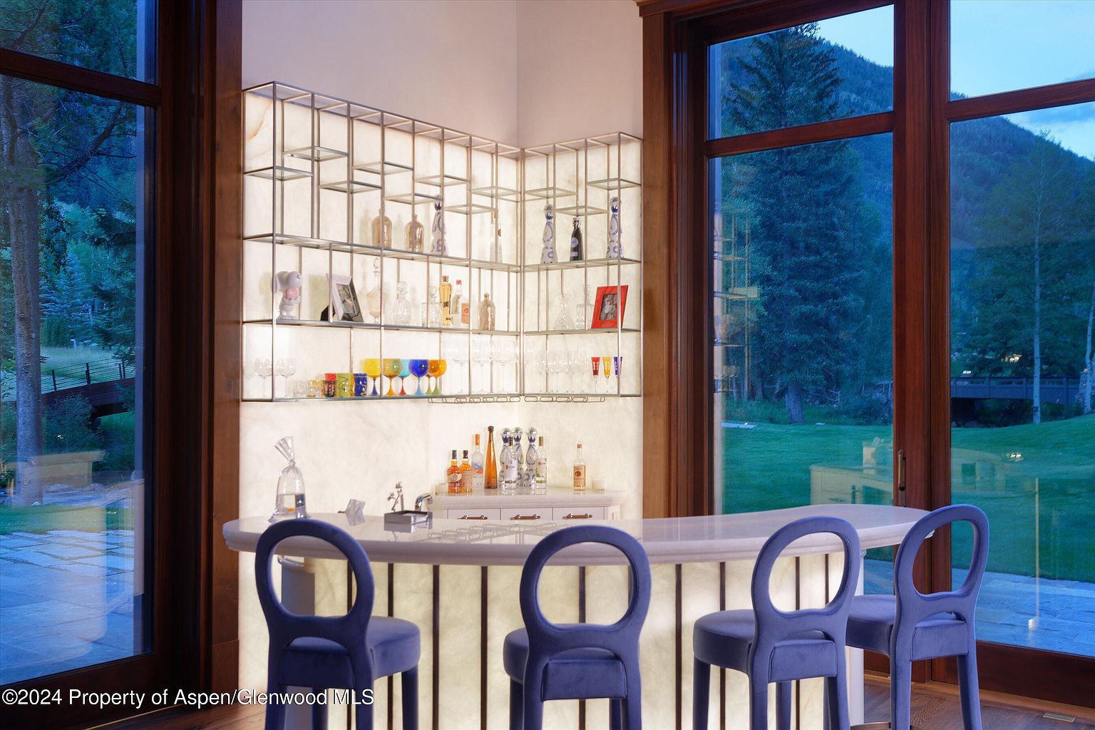This image showcases a stylish home bar area, featuring a backlit bar with a curved countertop and four blue velvet bar stools. The bar is equipped with open shelving displaying various bottles and glassware, illuminated by soft lighting. Large windows offer a view of the outdoor landscape, creating an inviting and sophisticated atmosphere.