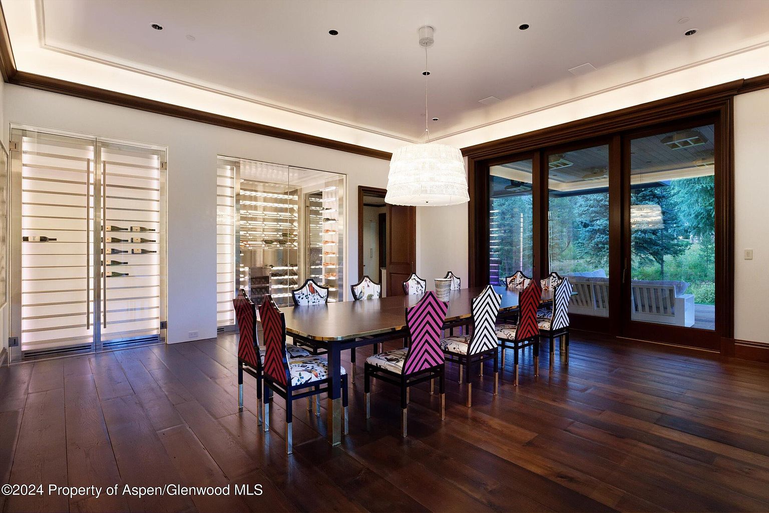 This dining room features a long, dark wood table surrounded by uniquely patterned chairs, set against a backdrop of hardwood floors and a large sliding glass door that offers a view of the outdoors. Two wine cellars flank the room, adding a touch of luxury and sophistication. A modern chandelier hangs above the table, providing ample lighting and enhancing the room's contemporary style.