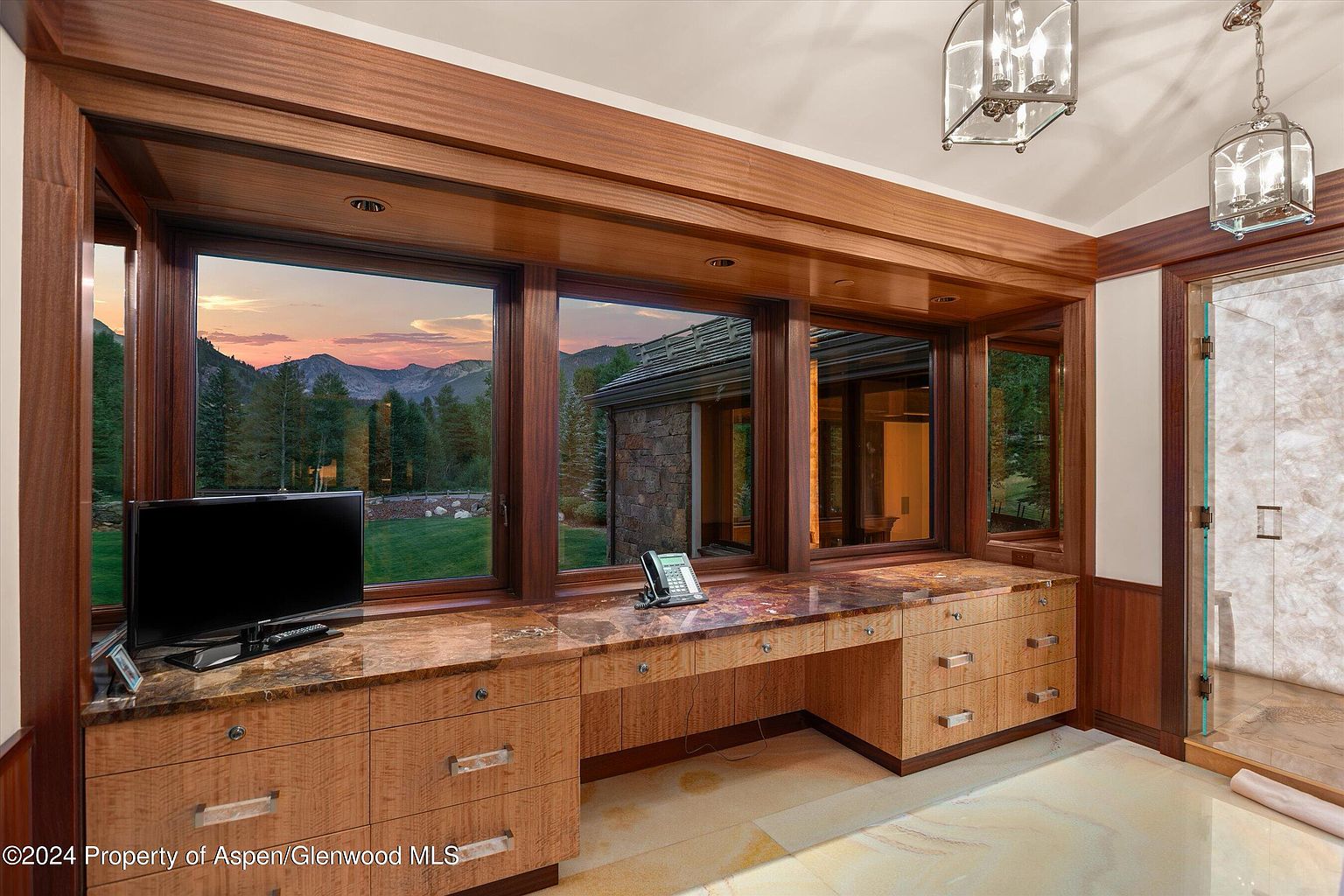 This is a luxurious primary bathroom featuring custom wood cabinetry with a marble countertop and a built-in desk area. Large windows offer a scenic view of the mountains and surrounding landscape. A glass-enclosed shower is visible to the right, and the overall design exudes elegance and sophistication.