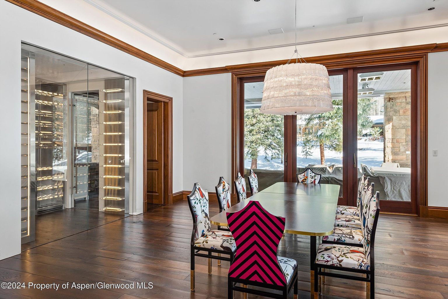 This dining room features a long, dark wood table surrounded by uniquely patterned chairs, with a striking pink and black chevron chair adding a pop of color. A large, modern chandelier hangs above the table, and expansive windows offer a view of a snowy landscape. A glass-enclosed wine cellar is visible to the left, adding a luxurious touch to the space.