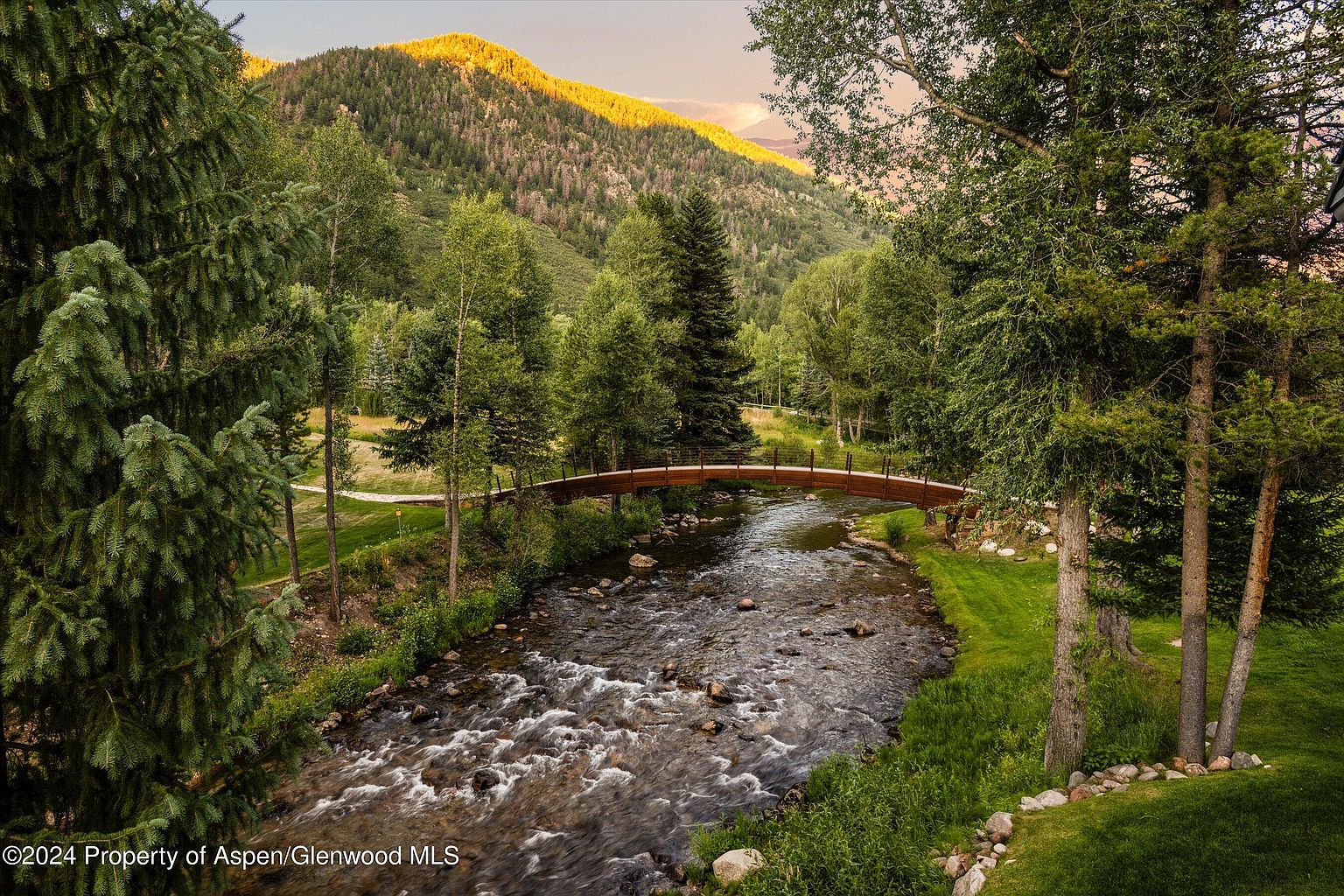This picturesque outdoor scene features a gently flowing river crossed by a charming wooden bridge, surrounded by lush greenery and mature trees. A mountain rises in the background, bathed in the warm light of the setting sun. The overall impression is one of tranquility and natural beauty, highlighting the property's serene and private setting.