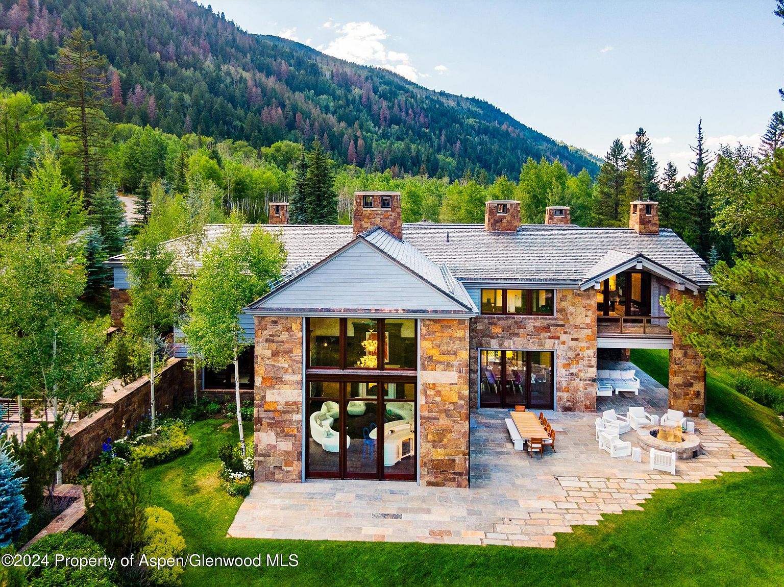 This is an aerial rear view of a luxurious stone house nestled in a lush, green landscape with a mountain backdrop. The house features a stone facade, a gray roof, multiple chimneys, and large windows. An outdoor patio area includes a dining table, chairs, a fire pit, and lounge seating, creating an inviting space for relaxation and entertainment.